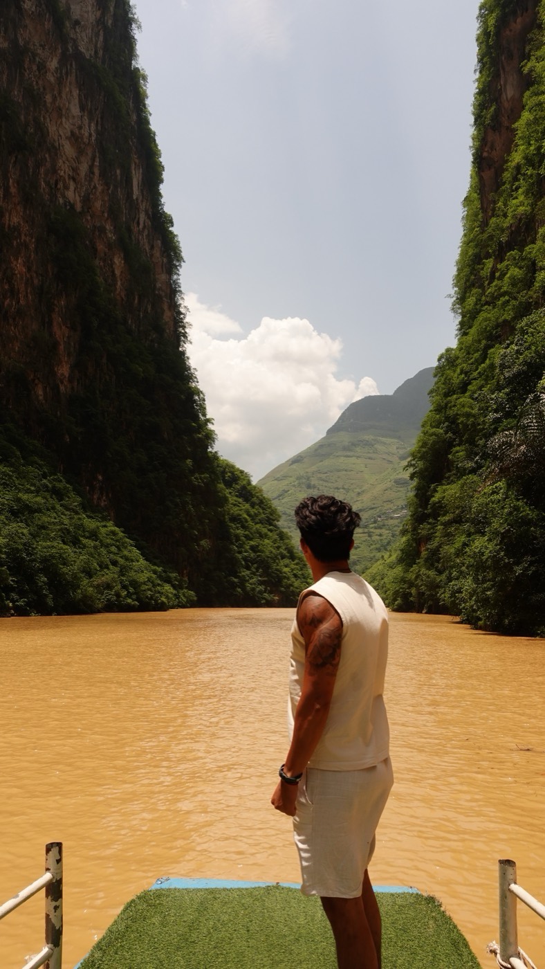 Standing at the front of a boat on a wide brown river, steep green cliffs rising on both sides